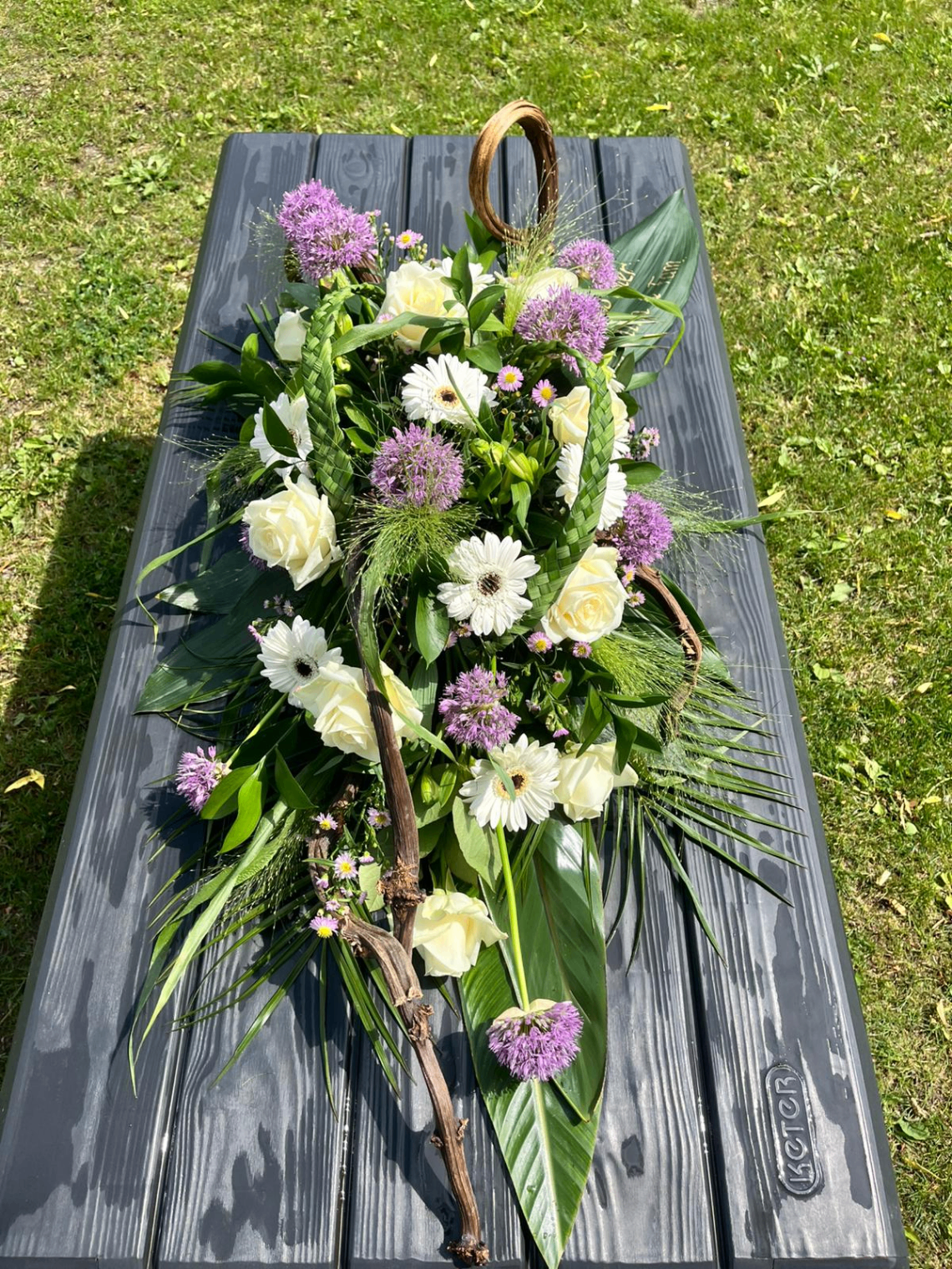 Langwerpig afscheidsbloemstuk met witte rozen, paarse alliums en gevlochten groen